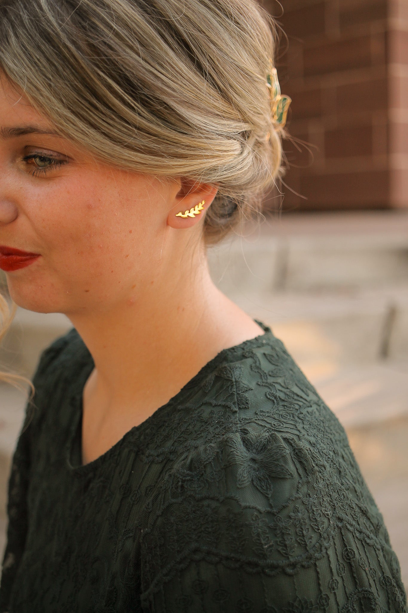 A young woman with blonde hair, styled elegantly, wears a gold leaf earring. She is dressed in a dark green, textured top, set against blurred stone steps in a natural light.