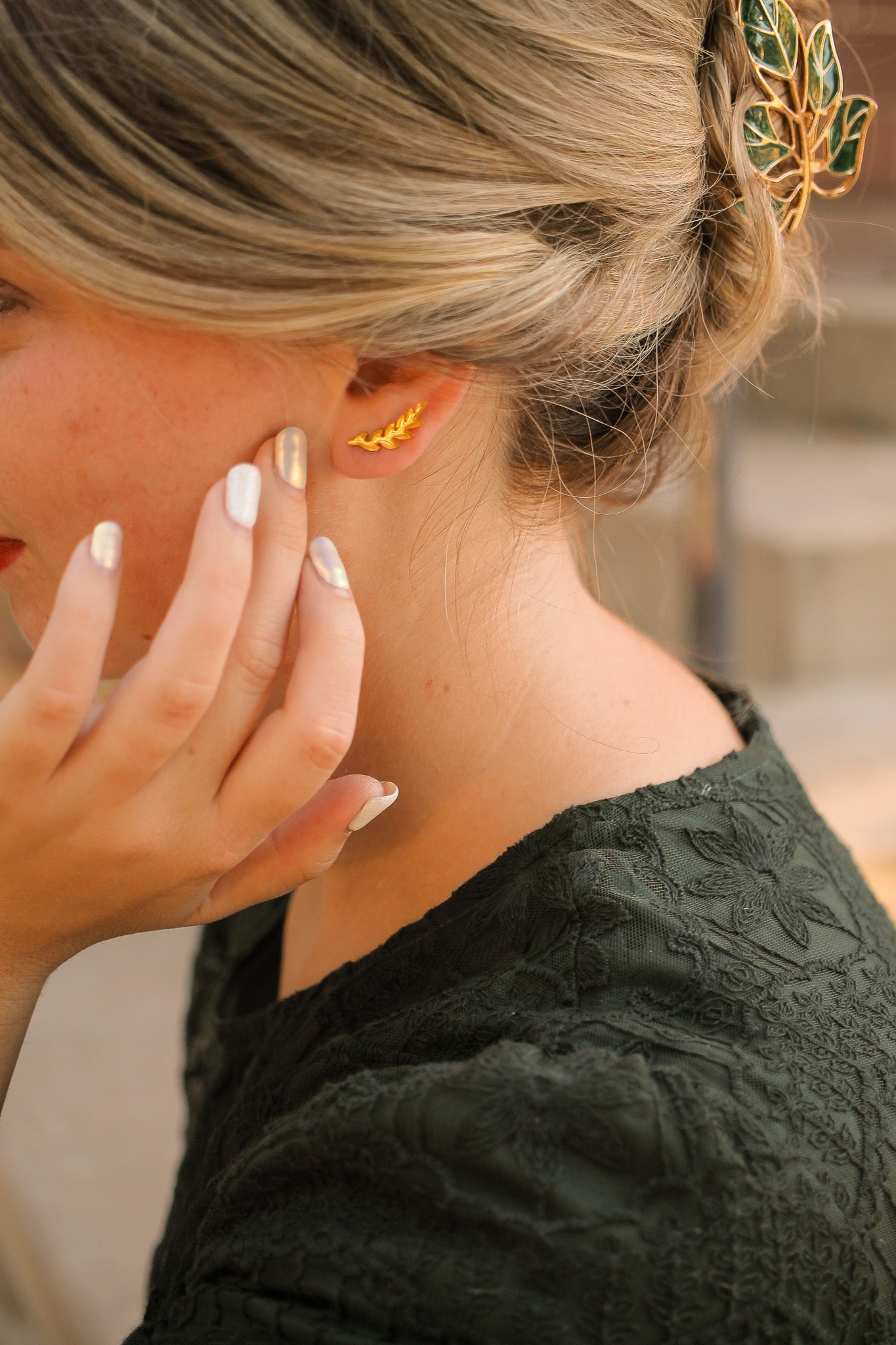 A woman with long, wavy hair touches her ear, showcasing a golden leaf-shaped earring. She wears a dark green textured top, set against a soft, blurred outdoor background.