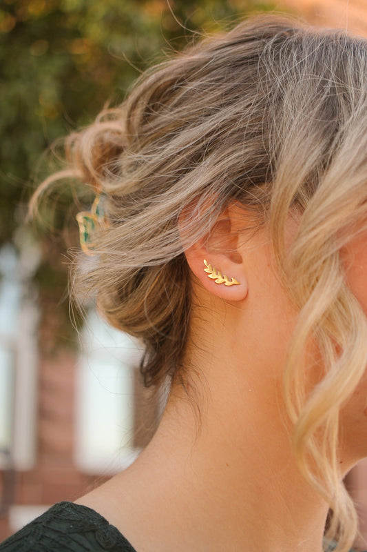 A womanâ€™s ear is adorned with a gold leaf-shaped earring. Her blonde hair is styled in loose waves, caught in natural light, against a blurred background of greenery.