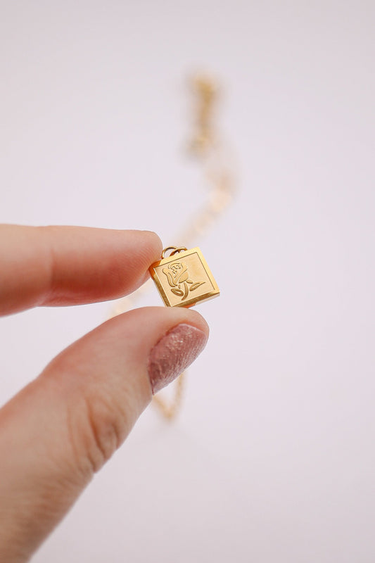 A person holds a small, square gold pendant engraved with a rose. The pendant is suspended near a delicate gold chain, set against a soft, light background.