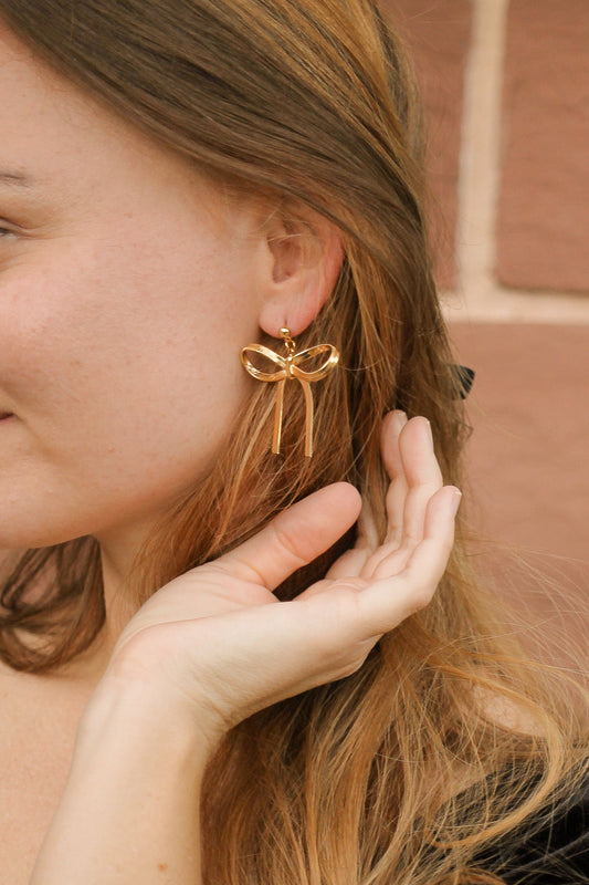 A woman is gently touching her hair while showcasing a large, gold bow-shaped earring on her ear. The background features textured walls, hinting at an outdoor setting.