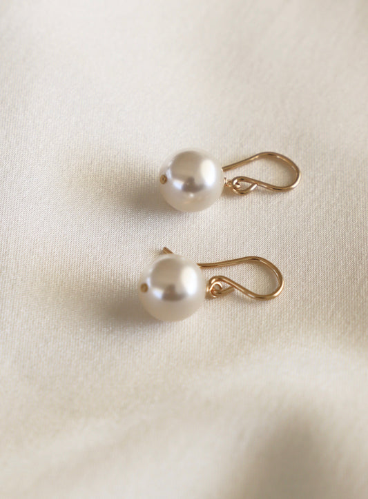 Two elegant pearl earrings hang on a soft, light-colored fabric. The pearls are round and glossy, with a delicate gold clasp that complements their appearance.