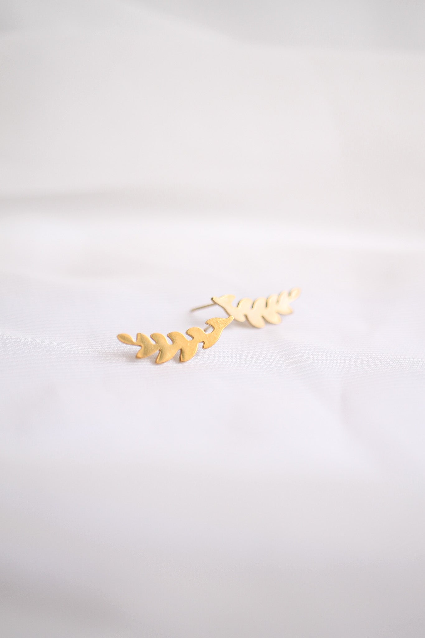 A pair of gold leaf-shaped earrings rests on a soft, white fabric background, showcasing the delicate design and shiny finish. They appear elegantly crafted and ready for wear.