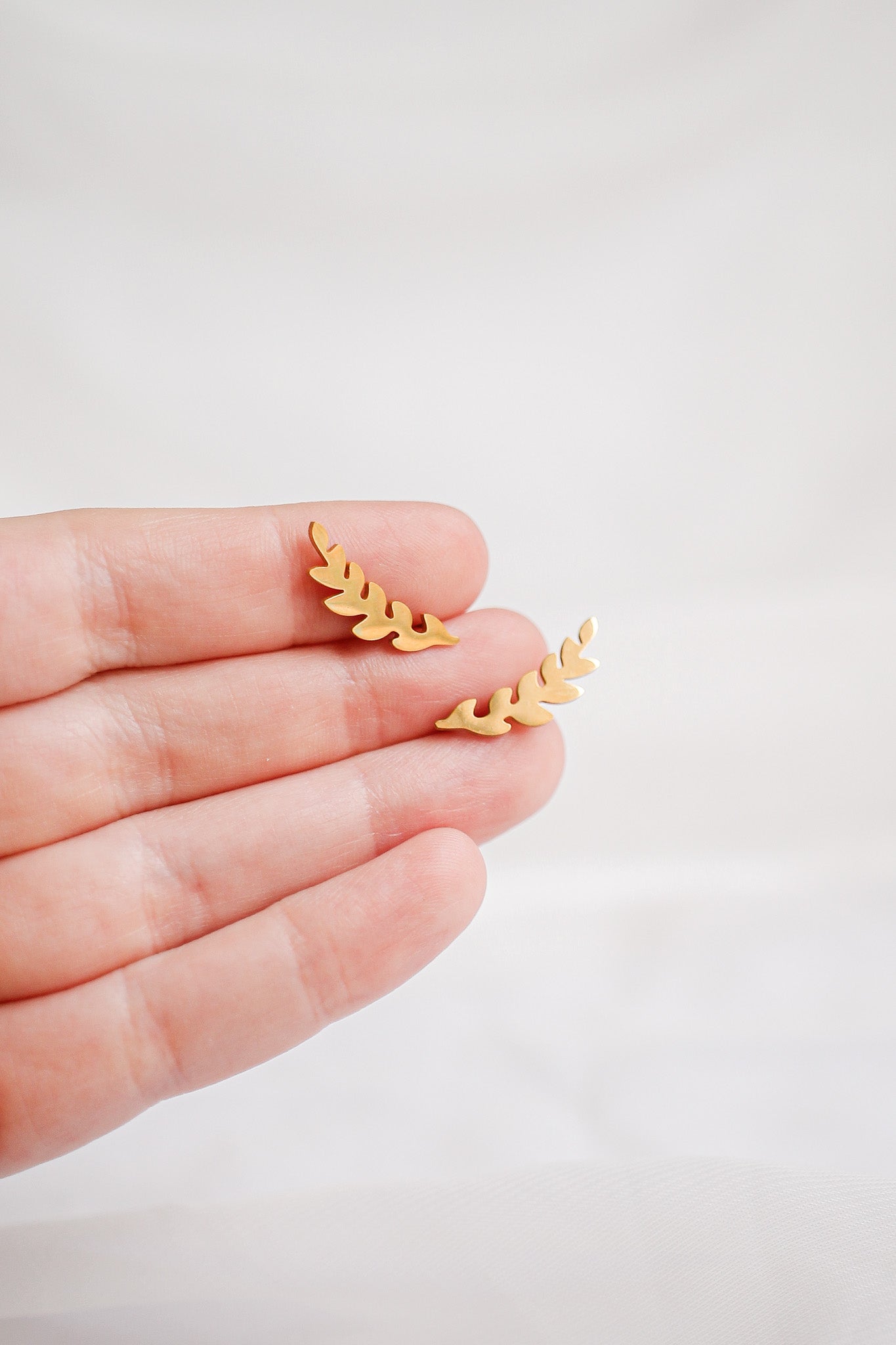 A hand holds two small, gold-colored leaf-shaped earrings, showcasing their intricate design against a soft, light-colored background that suggests a gentle and elegant atmosphere.