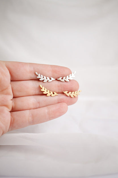 A hand holds two pairs of decorative leaf-shaped earrings, one pair in white and the other in gold, against a soft, neutral-colored background.