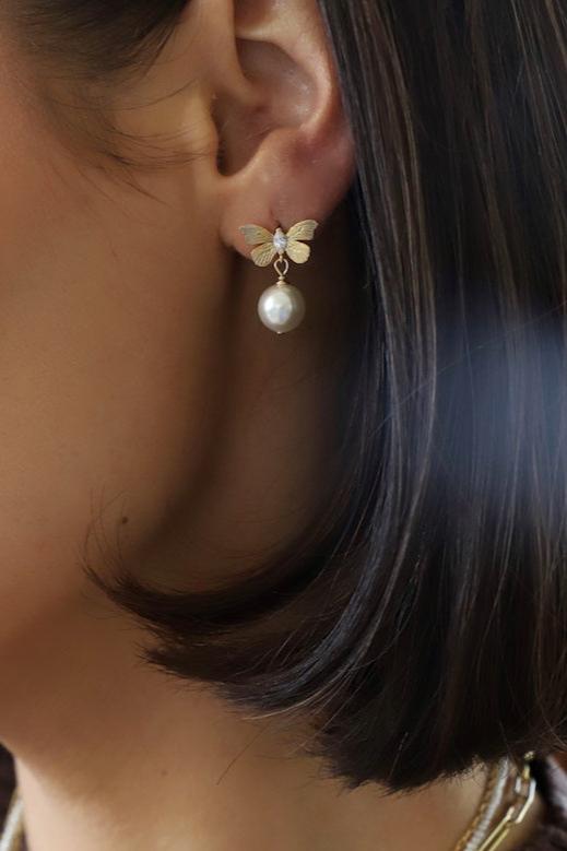 A butterfly-shaped earring adorned with a pearl is displayed on the ear of a person with short, dark hair. The setting is softly lit, suggesting a stylish atmosphere.