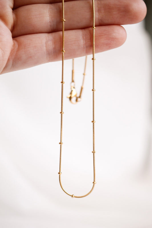 A delicate gold necklace is held up by a hand, displaying its fine chain and bead accents against a soft, neutral background, emphasizing its elegance and simplicity.