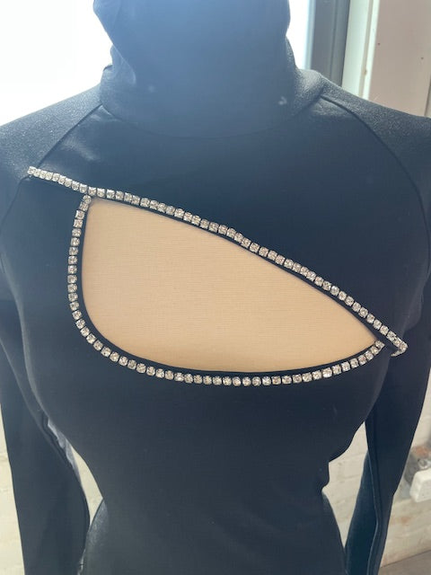 A black garment features a prominent cut-out section outlined with sparkling rhinestones, showcasing the underlying skin color. The fabric has a sleek texture and a high collar.