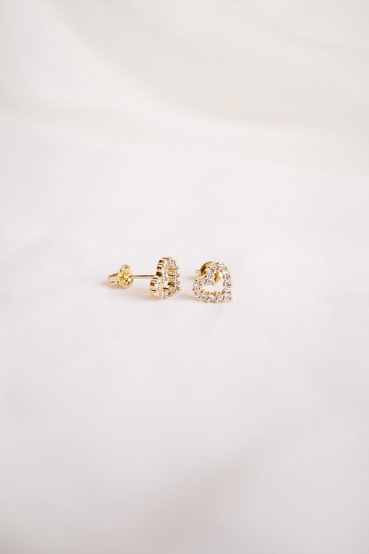 Gold heart-shaped earrings adorned with small sparkling stones lay on a soft white fabric surface, emphasizing their shine and delicate design in a minimalistic setting.