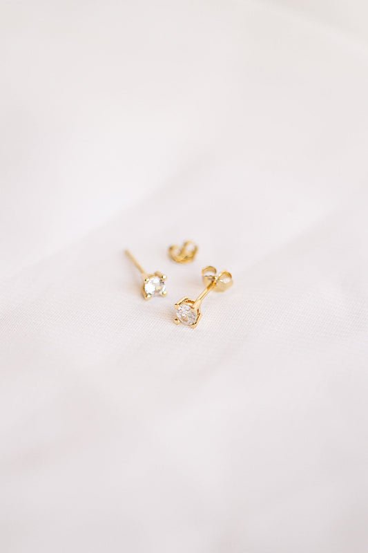 Three pairs of gold earrings with sparkling stones lie on a soft, light-colored fabric, showcasing their elegance and brilliance in a minimalistic setting.