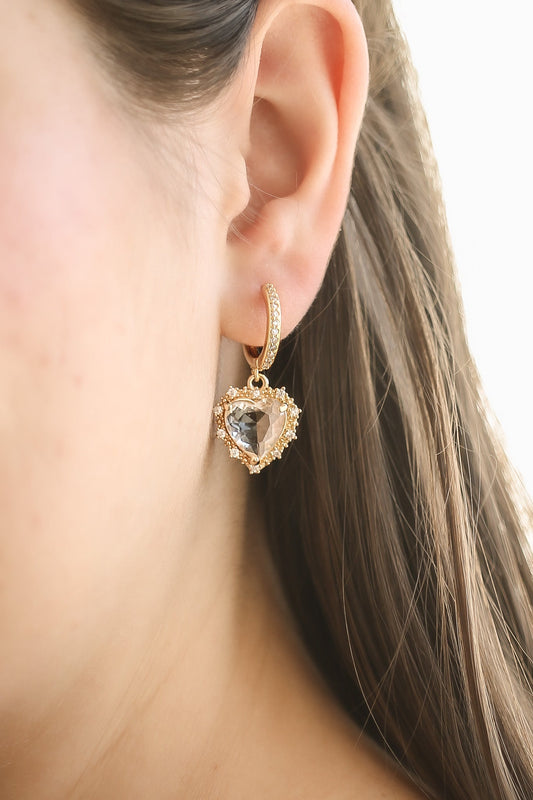 A close-up of a gold heart-shaped earring hangs from a woman's pierced ear, featuring a clear center stone surrounded by small decorative elements, with long brown hair partially visible.