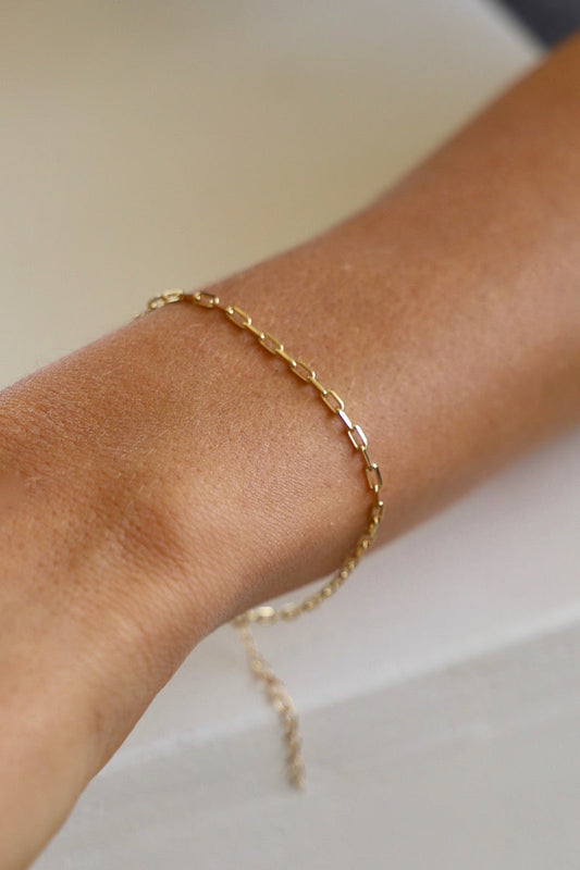 A slender gold chain bracelet adorns a tanned wrist, resting against a light-colored surface. The bracelet features interconnected rectangular links and a delicate chain extending from it.