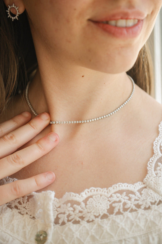 A woman is gently touching a sparkling silver necklace around her neck, showcasing its delicate design. She wears a white lace top and has long hair flowing beside her.