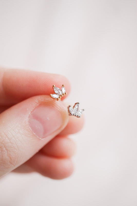 Two small earrings, one gold and one silver, shaped like crowns with multiple clear gemstones, are held between the thumb and index finger against a light, blurred background.