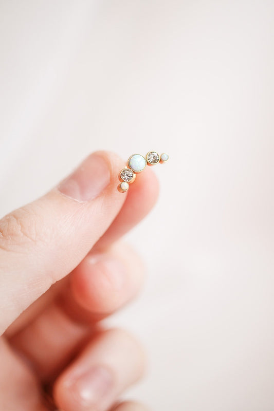 A hand holds a delicate piece of jewelry featuring three stones: a central opal and two adjacent gemstones. The background is neutral and softly blurred, emphasizing the jewelry.