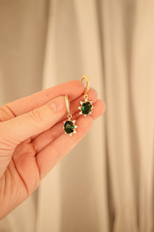 A hand holds two elegant earrings featuring green gemstones surrounded by sparkling accents, displayed against a soft, neutral-colored background that suggests a decorative setting.