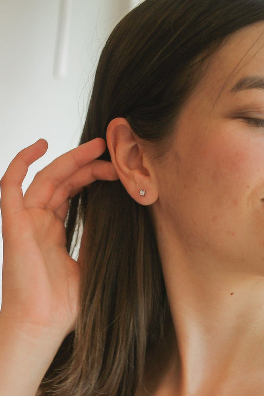 A woman gently touches her ear, highlighting a small, sparkly earring. Her long hair falls beside her face, and soft lighting creates a warm indoor atmosphere.