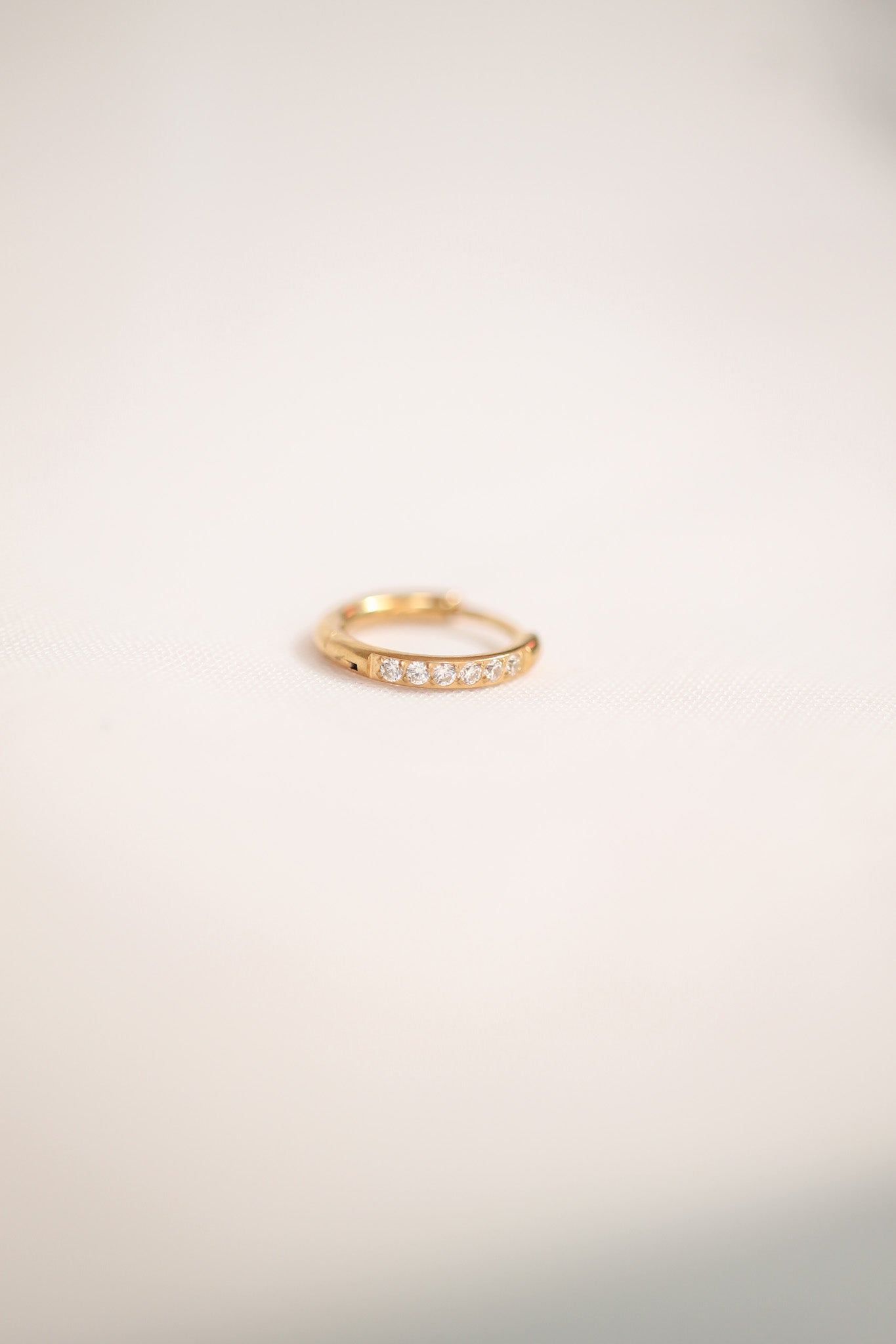 A gold ring with small diamonds is placed on a soft, neutral-colored fabric. The ring features a smooth design and shines softly, adding elegance to the minimalistic setting.