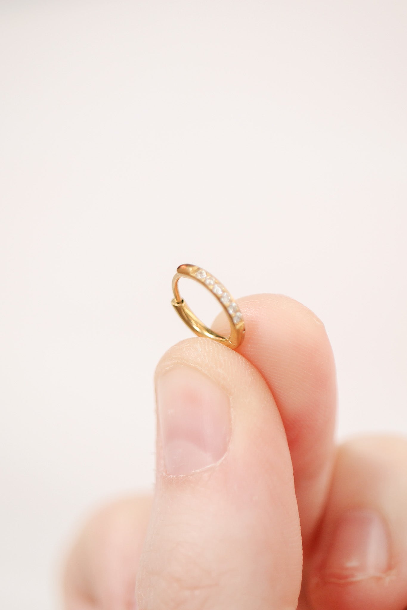 A person holds a small gold hoop earring with a row of tiny, clear gemstones. The background is a soft, pale color, enhancing the earring's features.