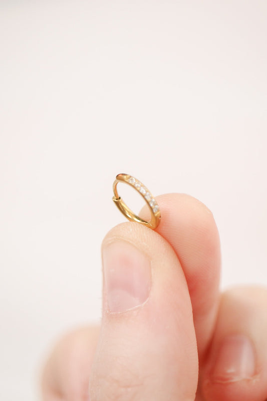 A person holds a small gold hoop earring with a row of tiny, clear gemstones. The background is a soft, pale color, enhancing the earring's features.