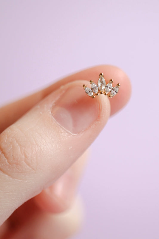 A hand holds a small, ornate piece of jewelry featuring multiple clear gemstones set in gold. The background is softly lit, with a light purple hue, enhancing the delicate appearance.