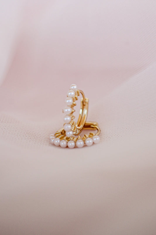 A pair of gold hoop earrings adorned with small pearls rests on a soft, pale pink fabric background, showcasing a delicate and elegant jewelry design in a serene context.