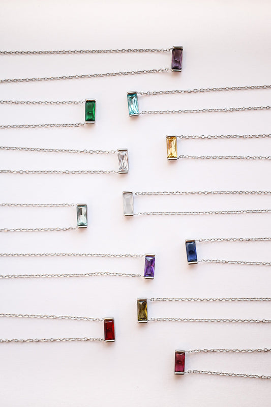 A collection of necklaces, each featuring a rectangular gemstone in various colors, suspended from thin silver chains, arranged neatly against a pale background.