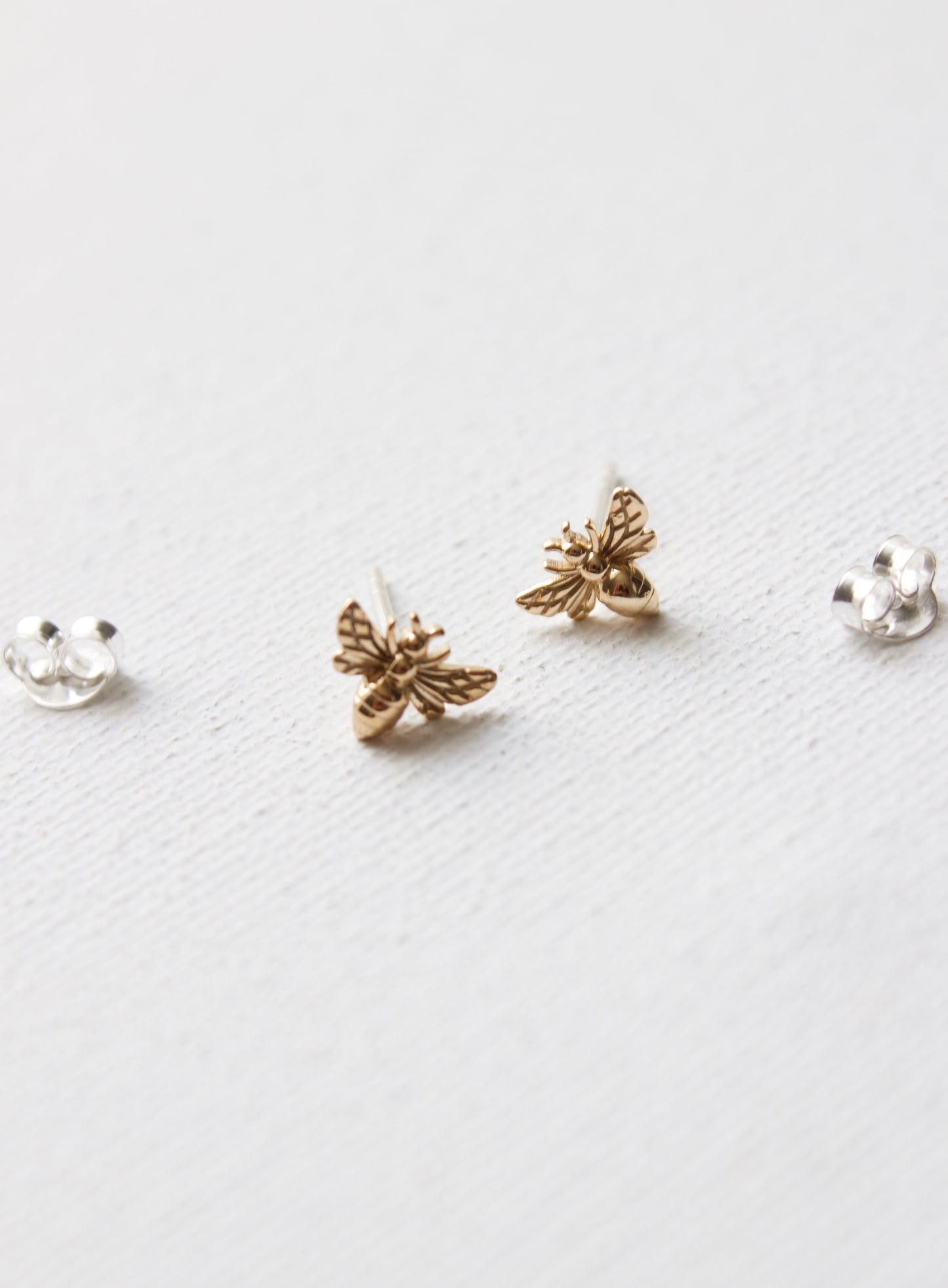 Two bee-shaped earrings, one gold and one silver, sit on a white textured surface. The earrings are side by side, showcasing intricate details of the bee design against the minimal background.