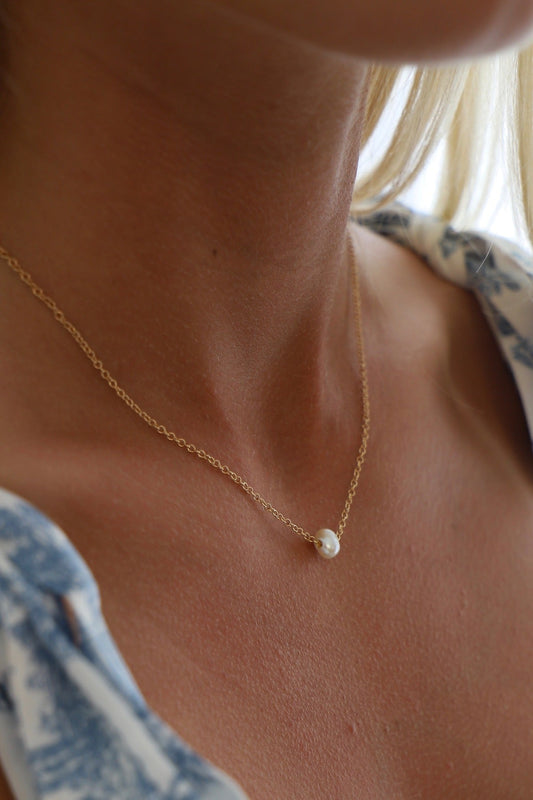 A delicate gold necklace with a single pearl rests on the skin of a neck, surrounded by soft light and hints of a patterned fabric in the background.