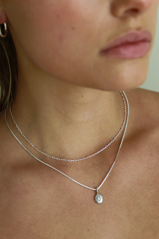 A close-up of a person's neck displays three silver chains, with the shortest featuring a decorative pendant. The skin tone is light, and the background is softly blurred.