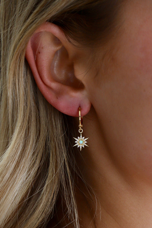 A gold hoop earring with a star-shaped pendant is hanging from a person's ear. The pendant features a central opal surrounded by small stones, set against a backdrop of long, light blonde hair.