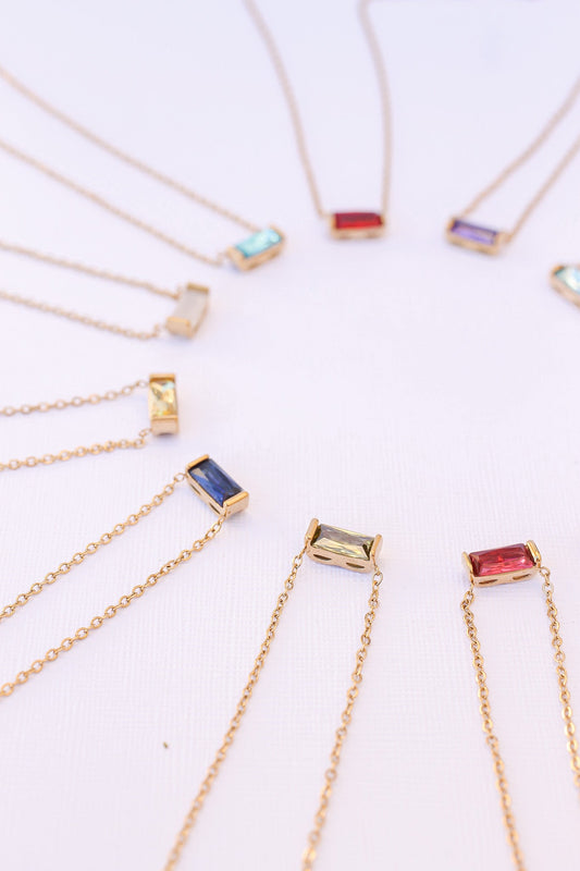 A circular arrangement of delicate gold necklaces with rectangular gemstone pendants in various colors, highlighting their elegance against a light background.