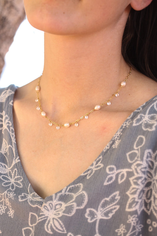 A delicate gold necklace with pearls and clear beads adorns a woman's neck, elegantly complementing her floral patterned blouse. The background hints at natural surroundings, soft and warm in tone.