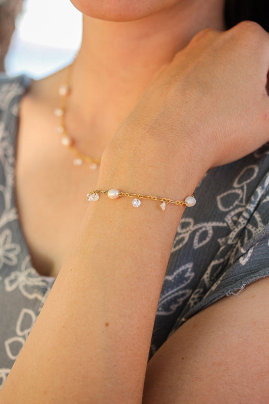 A delicate gold bracelet adorned with pearls and crystals is worn on a person's wrist. The setting features soft, natural light and a blurred, patterned fabric backdrop.