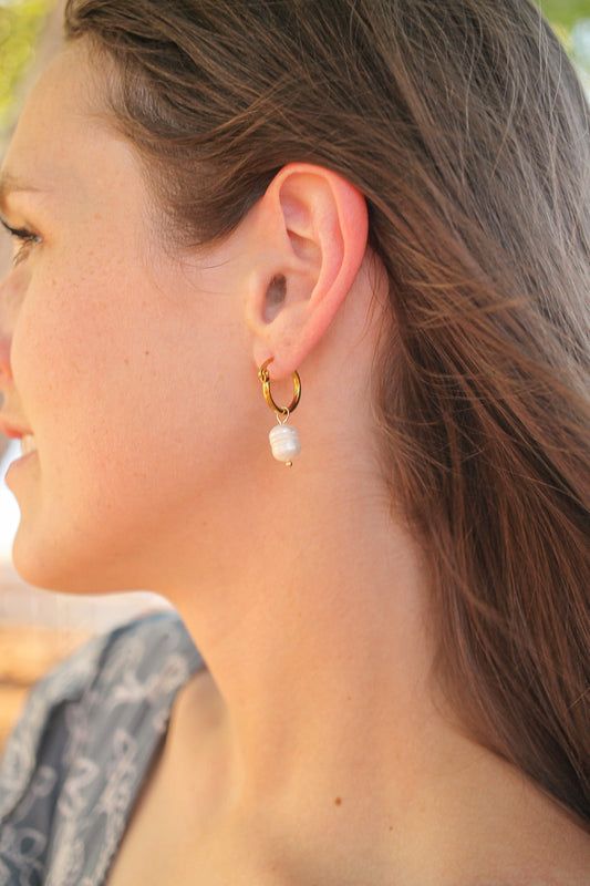 A womanâ€™s ear is adorned with a gold hoop earring featuring a white pearl. Her hair gently flows in a light breeze, creating a soft, serene atmosphere.