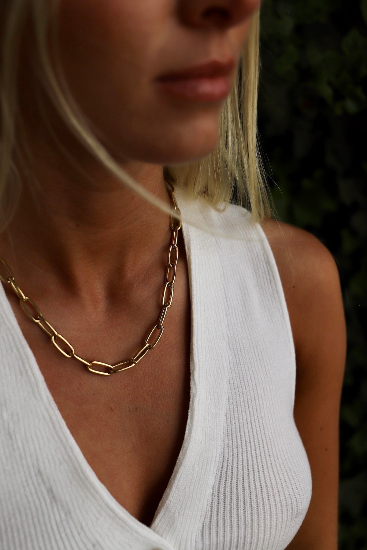 A gold chain necklace rests on a person's neck, contrasting against their textured white clothing. The background appears to be a blurred green foliage, enhancing the focus on the necklace.