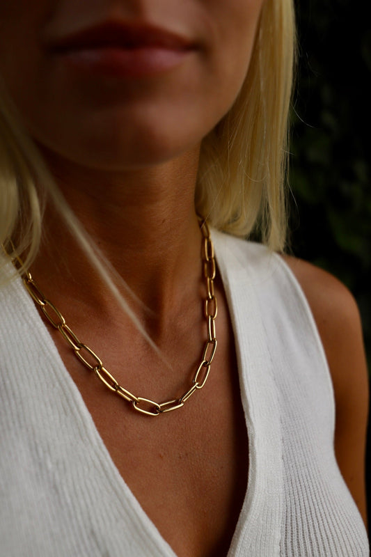 A gold chain necklace rests on the neck of a woman wearing a white, ribbed top, surrounded by green foliage, with her lips slightly visible in the frame.