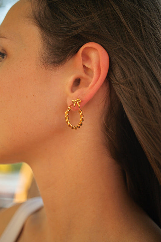 A gold hoop earring hangs from a womanâ€™s ear, showcasing a twisted design. Hair cascades down her shoulder, and a soft outdoor light creates a warm ambiance.