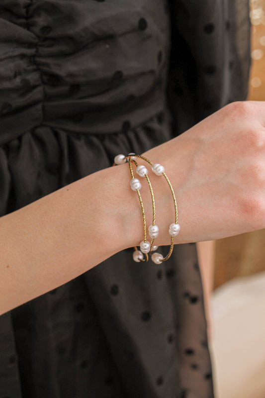 A woman's wrist adorned with a gold bracelet featuring multiple pearls. The wrist is actively displayed, surrounded by soft fabric of a black dress with textured polka dots.