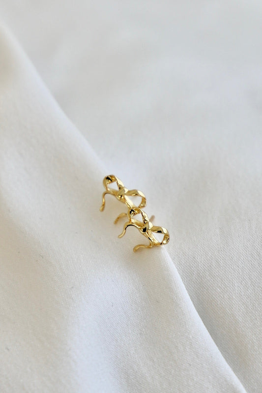 A pair of gold earrings rests on softly folded white fabric, showcasing a delicate design with intertwined shapes and curved edges, highlighting their elegant style in a minimalist setting.