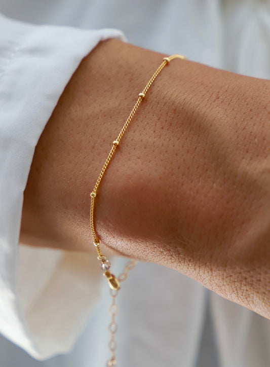 A delicate gold bracelet is worn on a tanned wrist, showcasing its fine chain and small embellishments. The background features part of a white shirt, softly blurred.