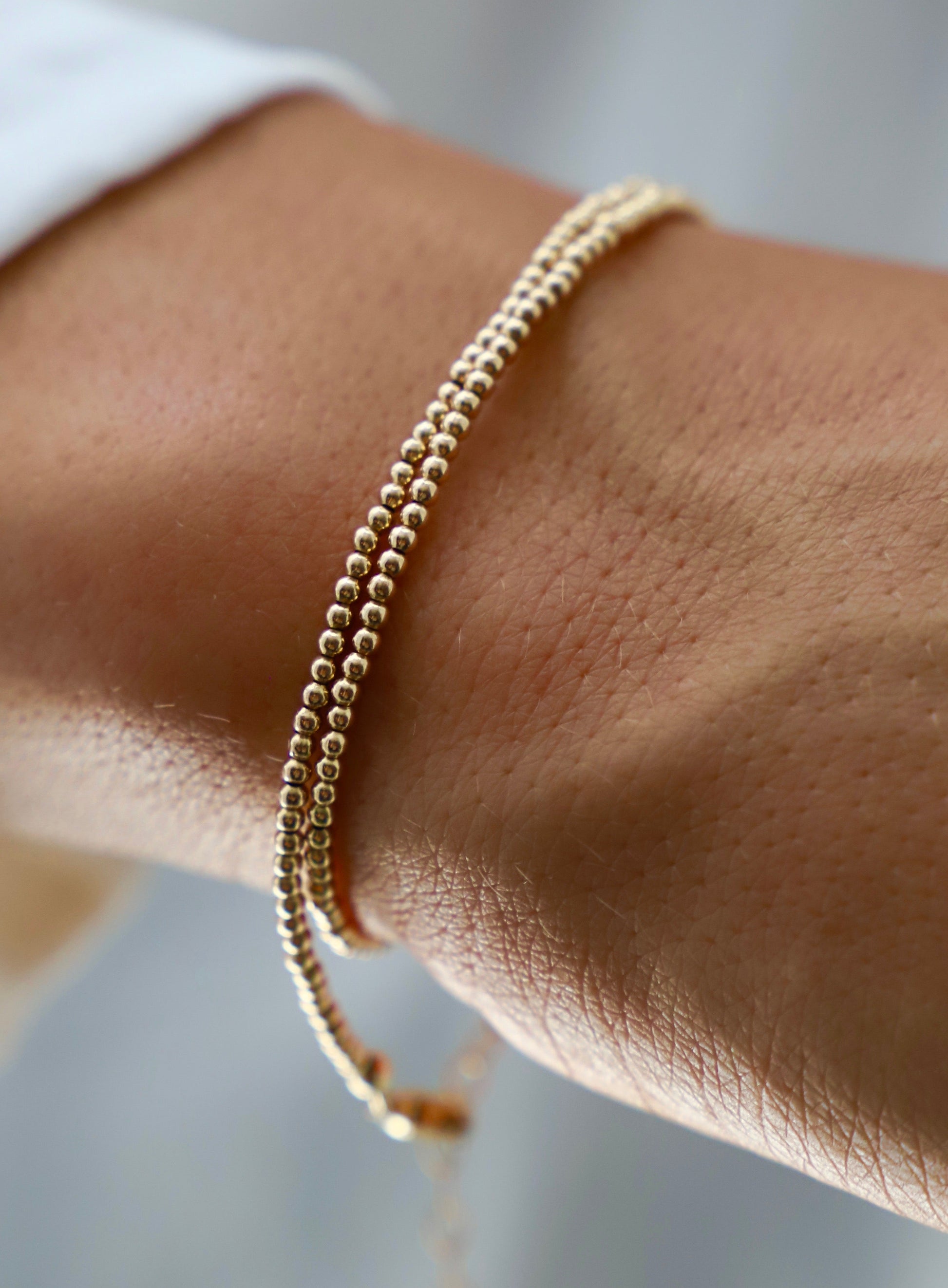 A gold bracelet with small spherical beads rests elegantly on a wrist, showcasing delicate skin texture. The background is softly blurred, creating a serene, focused atmosphere.