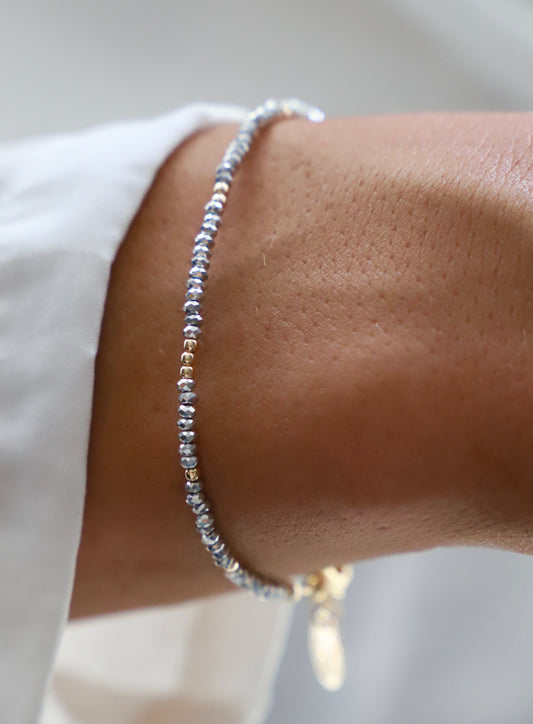 A delicate bracelet featuring alternating blue and gold beads rests on a bare wrist. The person's arm is partially visible, set against a light background with soft lighting.