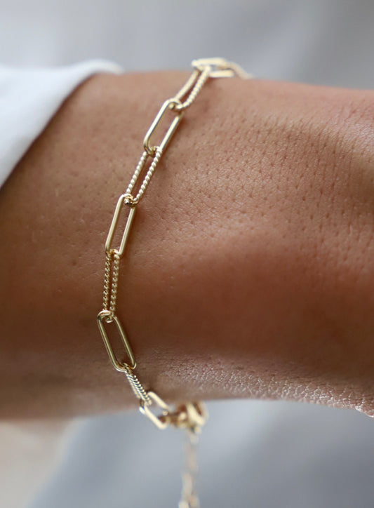 A gold chain bracelet wraps around a wrist, showcasing elongated links and a delicate design. The skin is visible, with a textured surface that contrasts with the shiny metal.