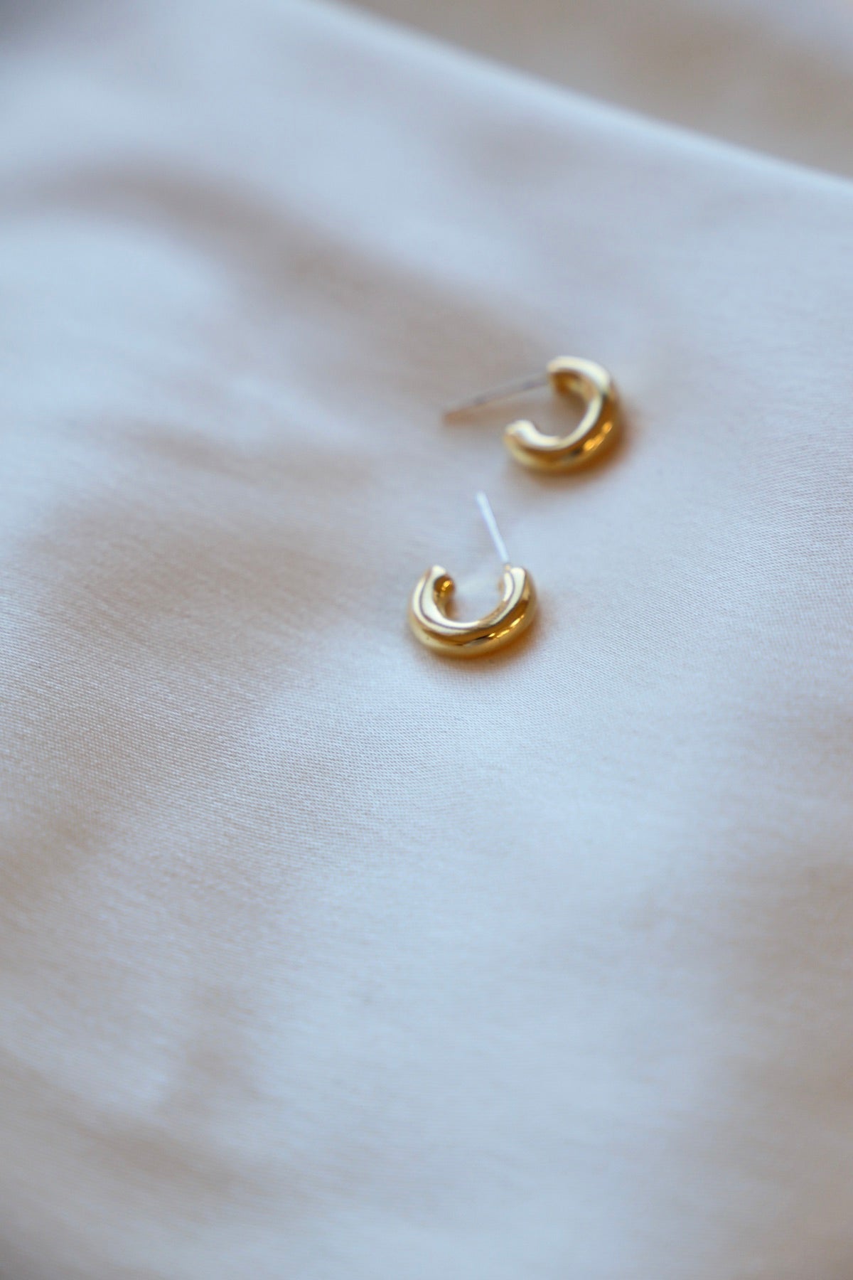 Two small, shiny gold hoop earrings rest on a smooth, light-colored fabric, suggesting a setting for jewelry display or preparation for wear.