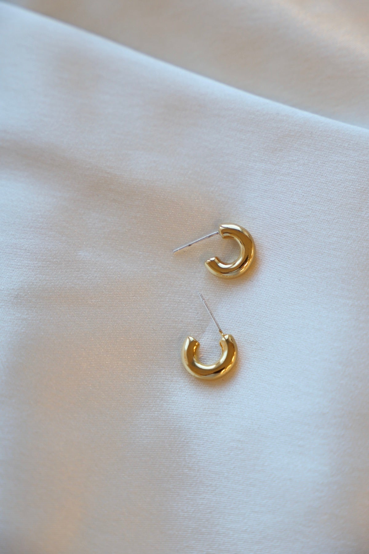 Two small gold hoop earrings rest on a smooth, light-colored fabric, reflecting soft light while appearing elegant and delicate in their design.