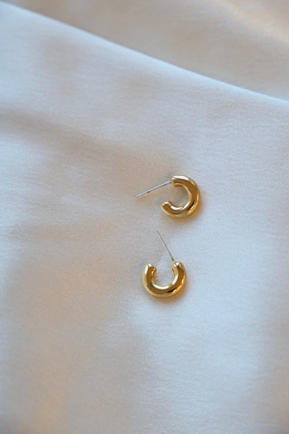 Two small gold hoop earrings rest on a smooth, light-colored fabric, reflecting soft light while appearing elegant and delicate in their design.