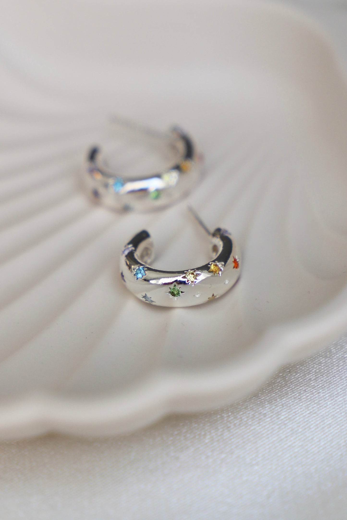 Two silver hoop earrings, adorned with colorful star-shaped gemstones, rest on a white, shell-shaped dish against a soft, light fabric background.