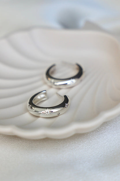 Two silver hoop earrings with star engravings rest elegantly on a white shell-shaped dish, set against a soft, light-colored fabric background.