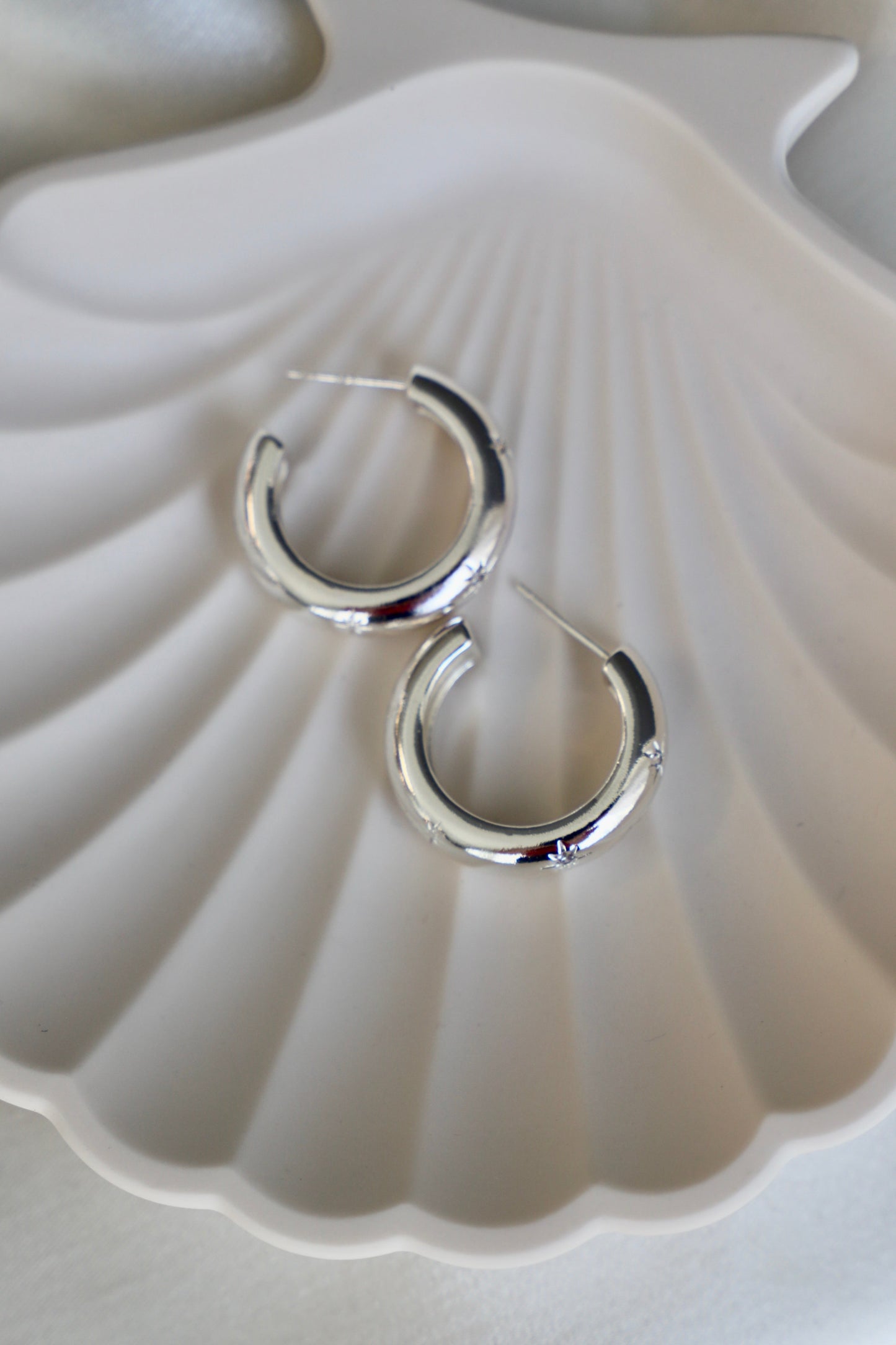 Silver hoop earrings rest on a white, scallop-shaped dish. The smooth surface reflects light, highlighting their shiny finish in a soft, neutral setting.